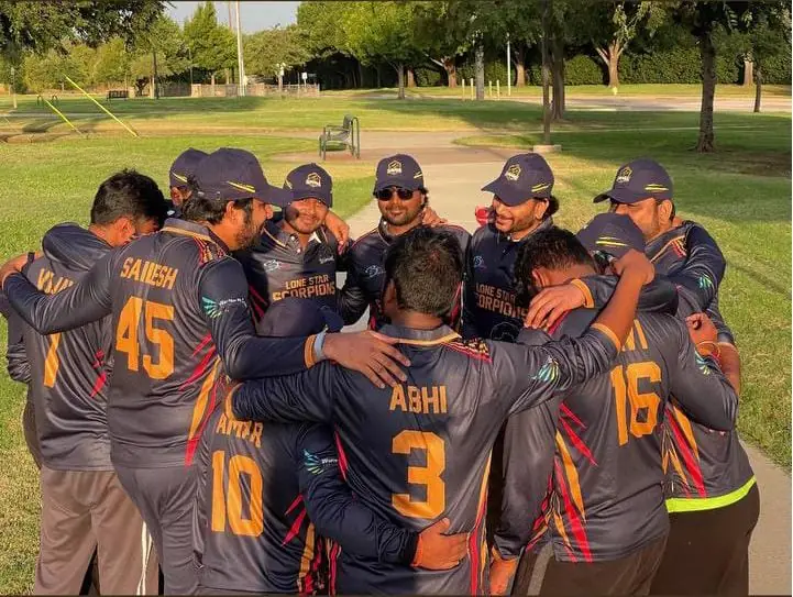 Lone Star Scorpions cricket team in a huddle wearing navy full-sleeve jerseys with orange names and numbers on the back and matching navy caps