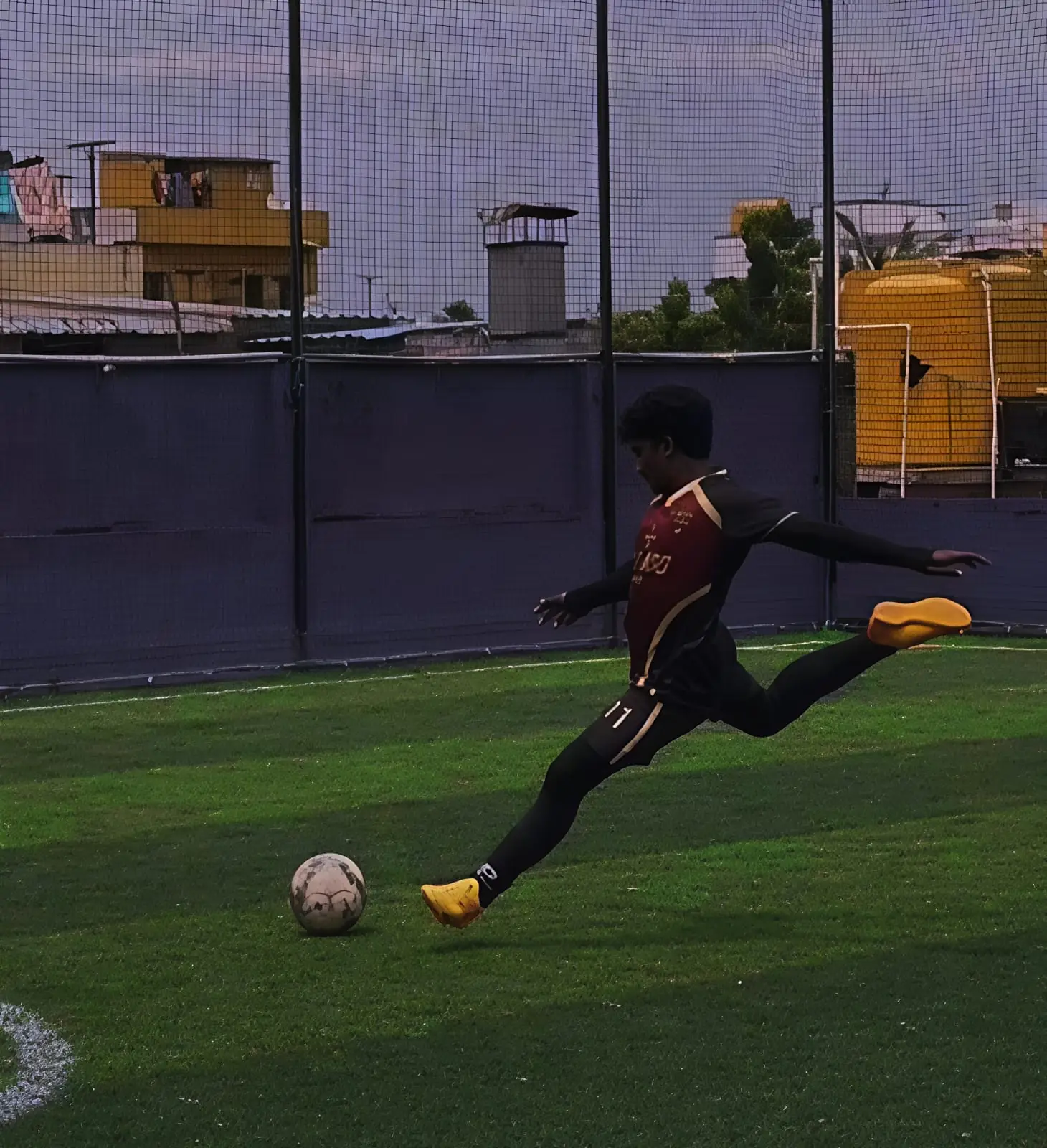 Footballer striking a ball on a caged turf in a maroon and black kit with gold trim and number 11 on the shorts